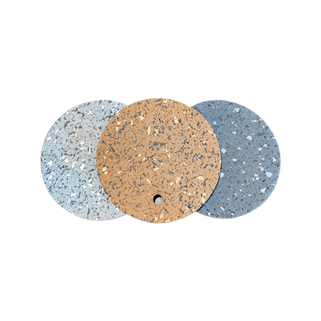 Three round samples in light grey, tan, and blue are arranged side by side on a white background, showing the speckled look of 5/16" (8mm) Kodiak Commercial Grade Rolled Rubber Flooring. The center tan sample has a small hole near its edge.