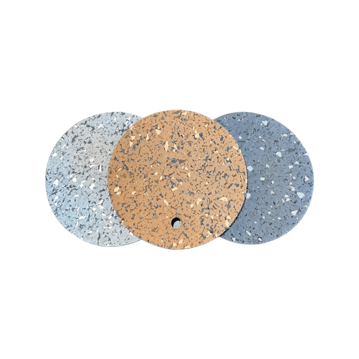 Three round samples in light grey, tan, and blue are arranged side by side on a white background, showing the speckled look of 5/16" (8mm) Kodiak Commercial Grade Rolled Rubber Flooring. The center tan sample has a small hole near its edge.